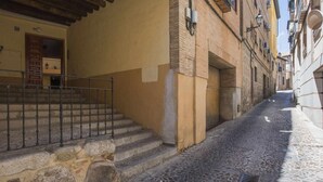 Un carrer empedrat i estret flanquejat per edificis d'arquitectura tradicional, una porta de fusta i una escala metàl·lica que condueix a un accés al segon pis.