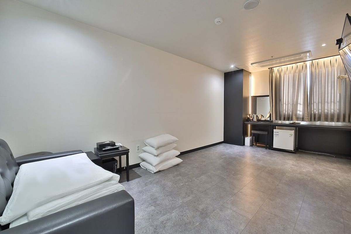 Una habitación de hotel moderna con un sofá gris, una mesita de noche pequeña, una lámpara, un espejo y una ventana con cortinas.