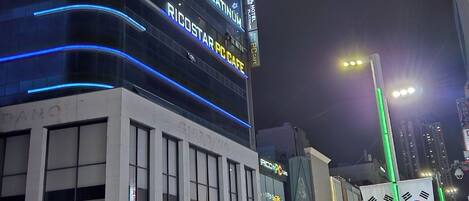 Een modern hotel met een gebogen gevel, borden met 'Hotel Platinum' en 'Ricostar PC Cafe', en een voetgangersgebied met straatverkopers en een rode scooter.