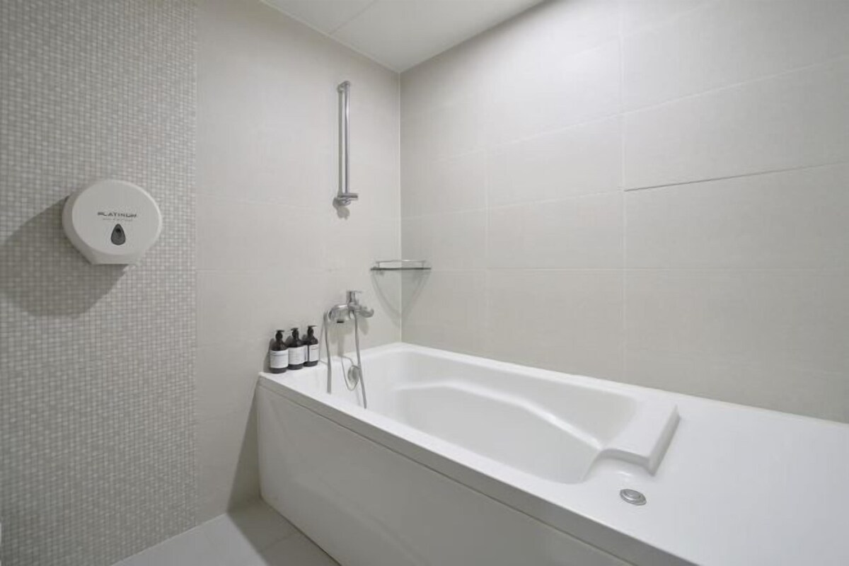 A modern bathroom with a white bathtub, a handheld showerhead, and a soap dispenser on the wall.