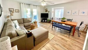 A living room with a sofa, ottoman, dining table, and chairs. There is a ceiling fan, a flat-screen TV, and framed artwork on the walls. - CapeSanBlas Bright, End Unit •Short walk to neighborhood private beach •2 Pools  (Port St. Joe)