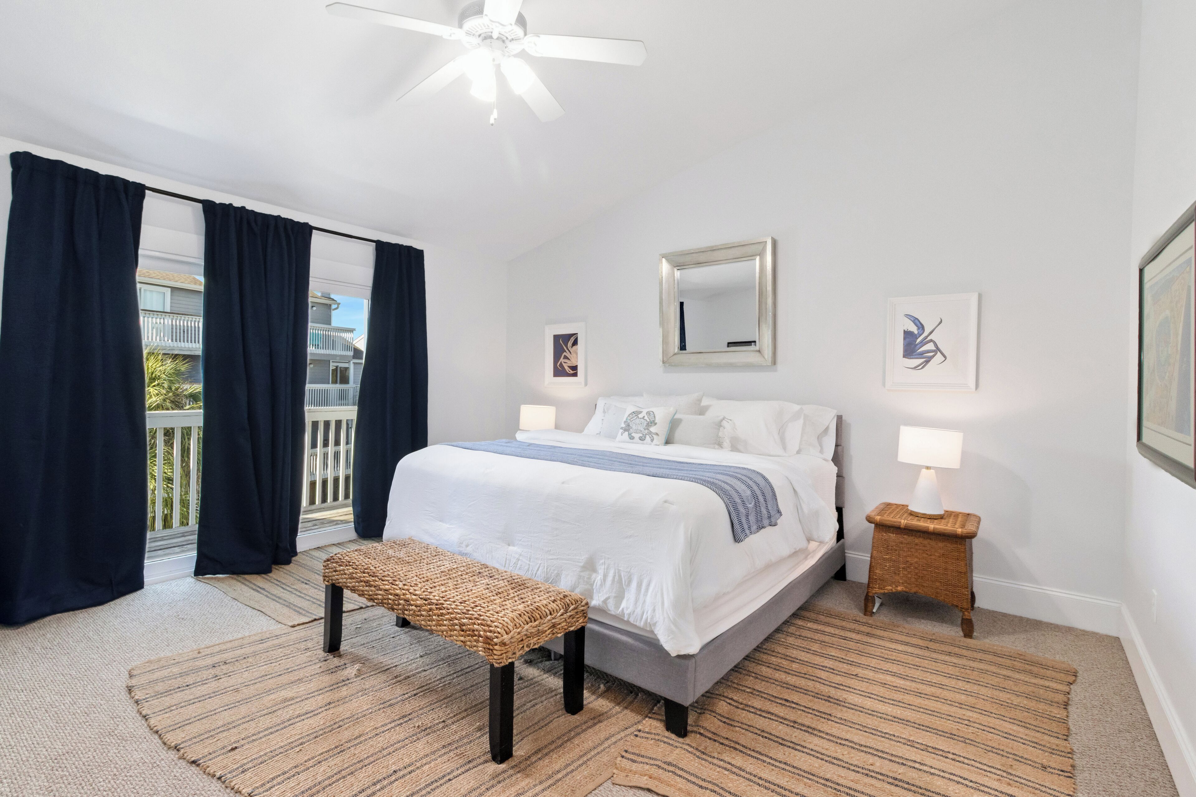 Une chambre à coucher comprenant un lit, un banc, une table de chevet avec une lampe, un ventilateur de plafond et un balcon avec des rideaux.