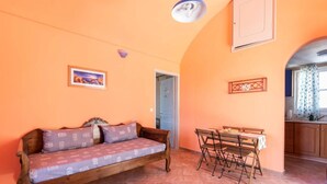 A cozy room with a wooden sofa, a dining table with chairs, a kitchen area, and a door leading to another room. - Santorious Nice View Apartment (Santorini)