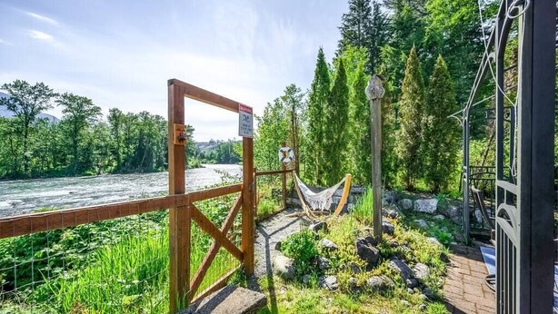 Ein hölzernes Tor führt zu einem Fluss, mit einer Hängematte und einer Holzhütte in der Nähe.