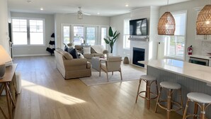 A spacious living room with a sofa, coffee table, and a TV mounted above a fireplace. There are wicker bar stools and a large window providing natural light. - Beautiful townhome in Wildwood Crest, walking distance to beach (Wildwood)
