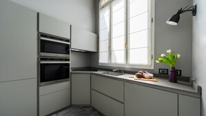 Une cuisine moderne avec des meubles de couleur grise, un four encastré, une fenêtre avec des stores blancs, un luminaire suspendu noir et une plante en pot posée sur le plan de travail.