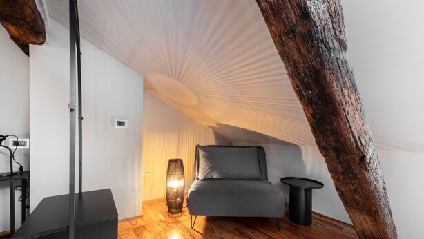 Une chambre à coucher confortable, aménagée en mezzanine, avec un plafond en pente, un plancher en bois, un canapé gris, une petite table ronde et un lampadaire.