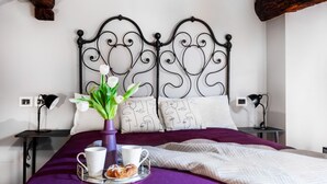 Un lit bien fait, recouvert d’une couvre-lit violette, avec une tête de lit décorative en métal, une table de chevet sur laquelle se trouve une lampe, et un plateau contenant une tasse, un mug et une pâtisserie.