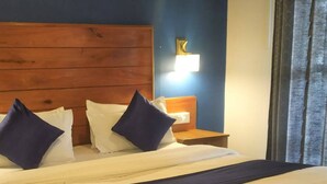 A hotel room with a wooden headboard, a bed with white and blue bedding, a nightstand with a lamp, and a view of the outside through a window with curtains. - Winter gardens mountain room (Munnar)
