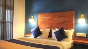 A hotel room with a wooden headboard, two bedside tables with lamps, a bed with white bedding and two dark blue pillows, and a blue accent wall. - Winter gardens mountain room (Munnar)