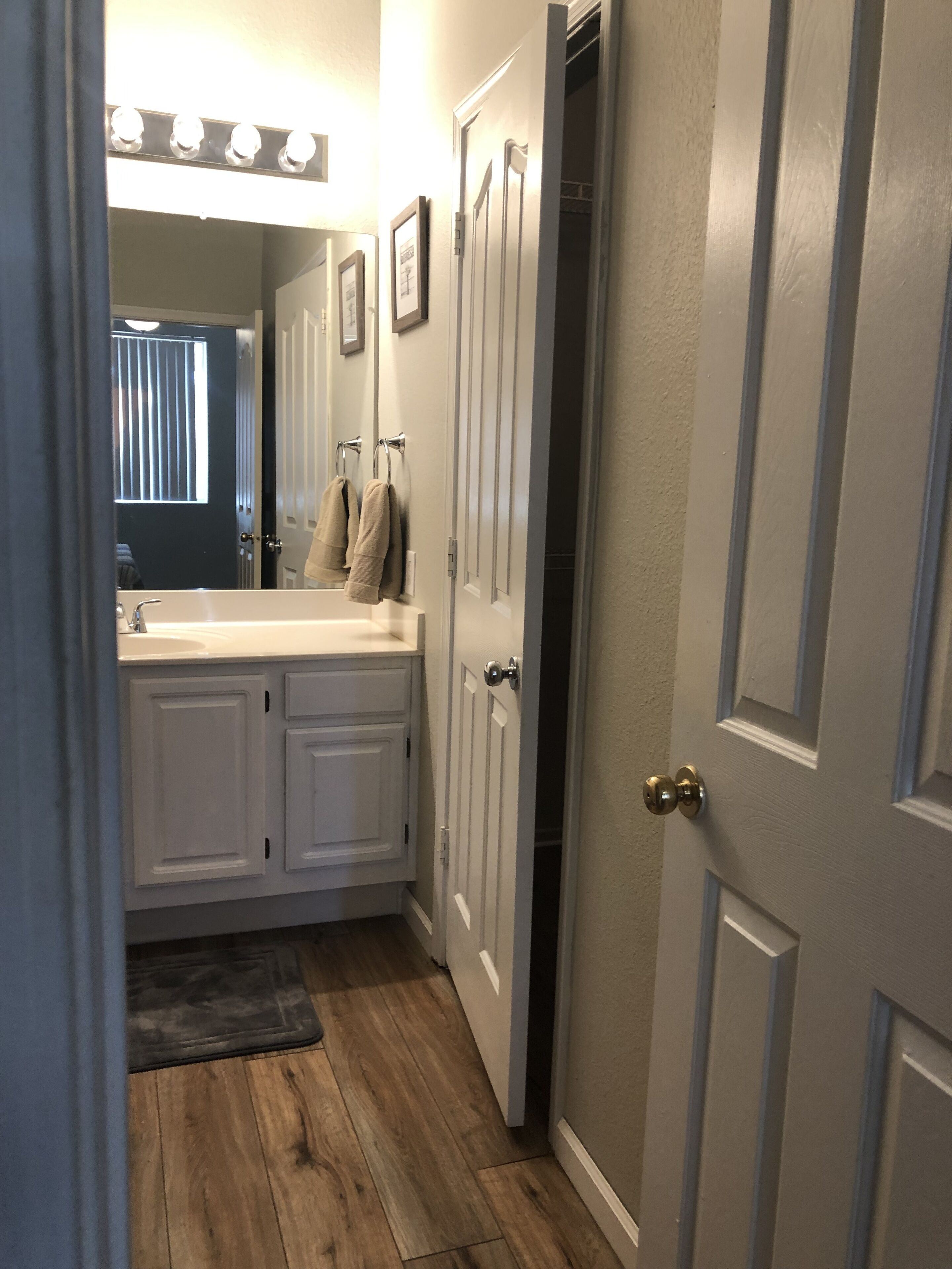 A bathroom with a vanity, sink, and mirror. There are towels hanging on the wall and a door leading to another room.