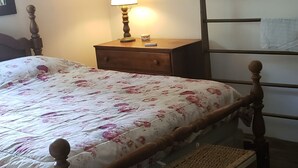 Een slaapkamer met een houten bed, een nachtkastje met een lamp, een slaapkamerkastje, een raam met gordijnen en een houten hoofdbord.