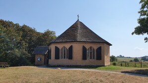 Une petite église coiffée d’un toit en forme de flèche et surmontée d’une croix, entourée d’un champ verdoyant et d’arbres.