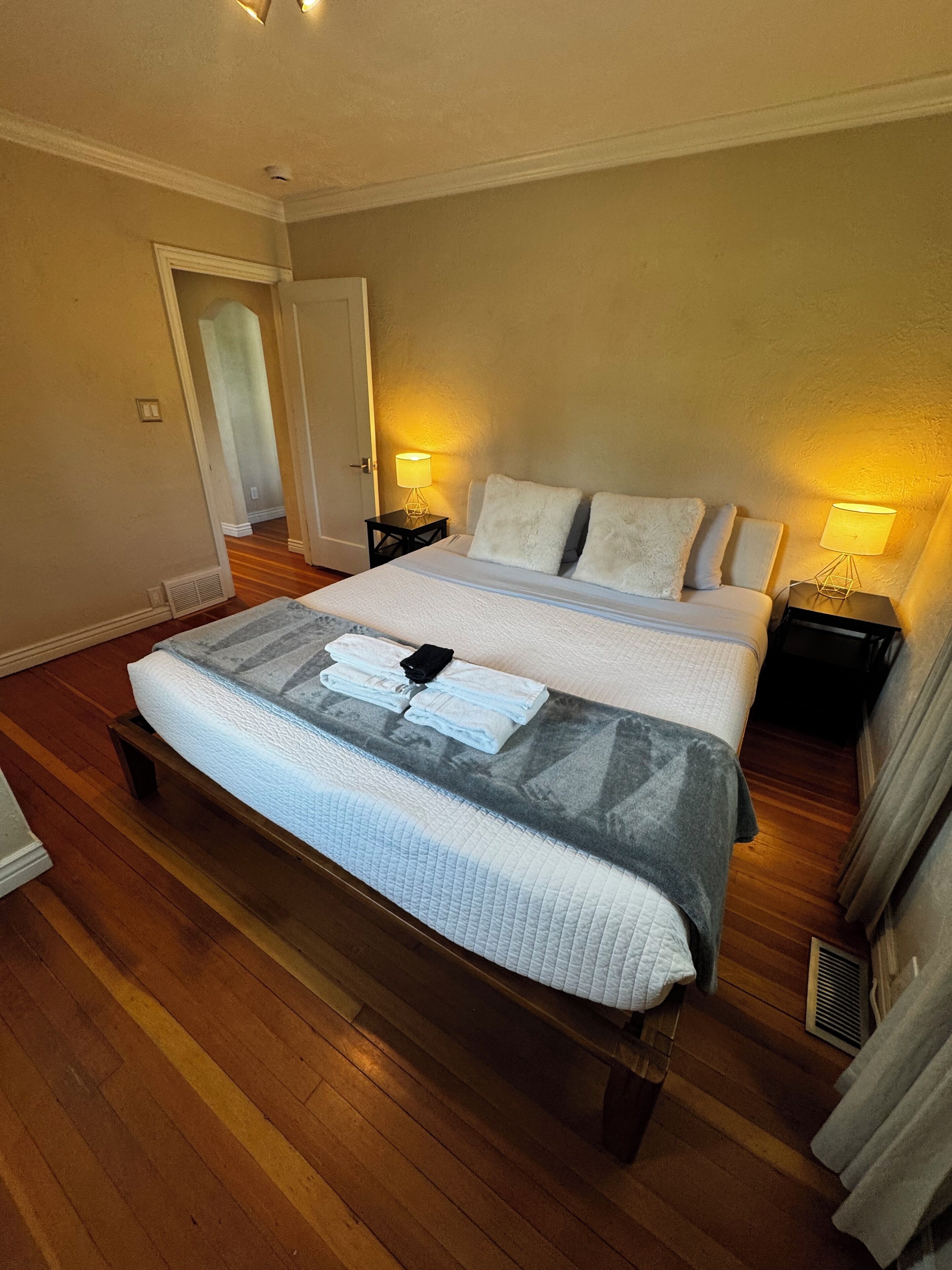A hotel room with a large bed, two bedside tables with lamps, wooden flooring, and a door leading to another room.