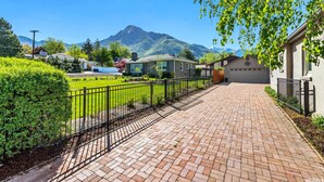 Une allée pavée mène à une maison avec un garage, qui est entourée d’une pelouse bien entretenue et, au loin, on aperçoit une montagne.