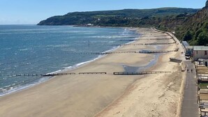 Une plage côtière avec une longue promenade, un ciel dégagé et des collines à l’horizon.