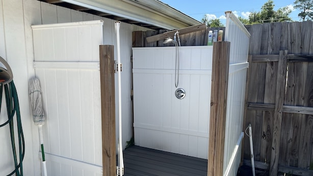 A small, enclosed outdoor shower area with a wooden deck, a white door, and a metal showerhead.