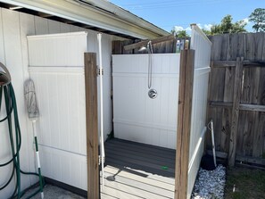 A small, enclosed outdoor shower area with a wooden deck, a white door, and a metal showerhead.