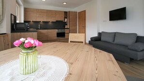Eine moderne Küche mit Holzschränken, einer Mikrowelle und einem Backofen. Ein Esstisch ist mit einer Blumenvase und einer Spitzendecke dekoriert. Im Wohnbereich steht ein graues Sofa mit einem Flachbildfernseher an der Wand.
