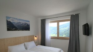 Ein Schlafzimmer mit einem hölzernen Kopfteil, einem Bett mit weißen Bettwäsche, einem Gemälde von Bergen, einem Fenster mit Vorhängen und einem angebrachten Fernseher.