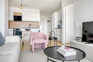 A modern living room with a sofa, a pink armchair, a round coffee table with books, a flat-screen TV, and a kitchen in the background.