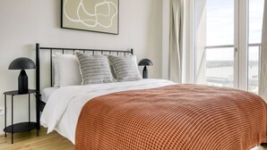 A modern bedroom with a large bed, two bedside tables with lamps, a view of the city through large windows, and a piece of abstract art on the wall. - 1BR w/ Balcony, Ideal view over Vienna, nr U2 Donaumarina, by Blueground (Vienna)