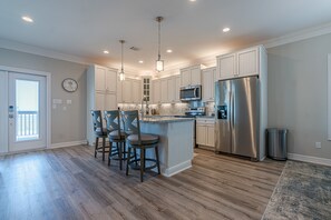 Una cocina moderna con piso de madera, muebles blancos, refrigerador de acero inoxidable e iluminación colgante.