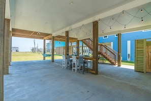 Un espacio de estar al aire libre con techo, con una mesa y sillas de madera, una escalera y vista a un edificio azul en el fondo.