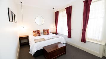 A bedroom with a bed, a bedside table, a wooden bench, a round mirror, and two windows with red curtains.