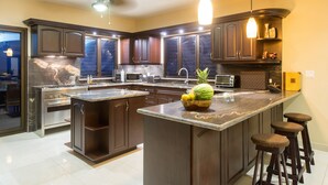 A modern kitchen with dark wood cabinets, a large island with stools, a microwave, and a toaster. - Luxury Oceanfront View Villa with Chef, Housekeeping and Concierge Services (Puerto Carrillo)