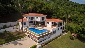 A two-story house with a swimming pool, surrounded by lush greenery and hills. - Luxury Oceanfront View Villa with Chef, Housekeeping and Concierge Services (Puerto Carrillo)