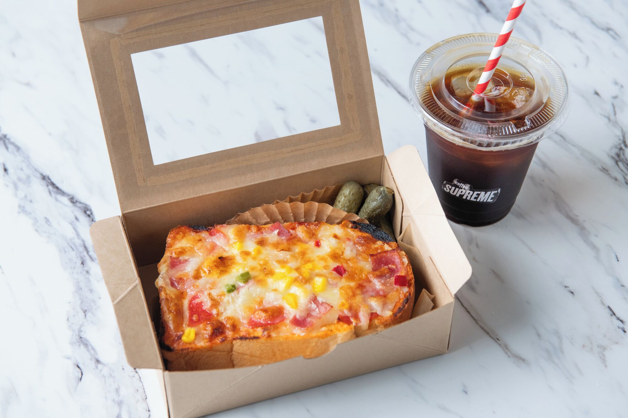 A cardboard box containing a slice of pizza with various toppings, a plastic cup of iced coffee, and two pickles.