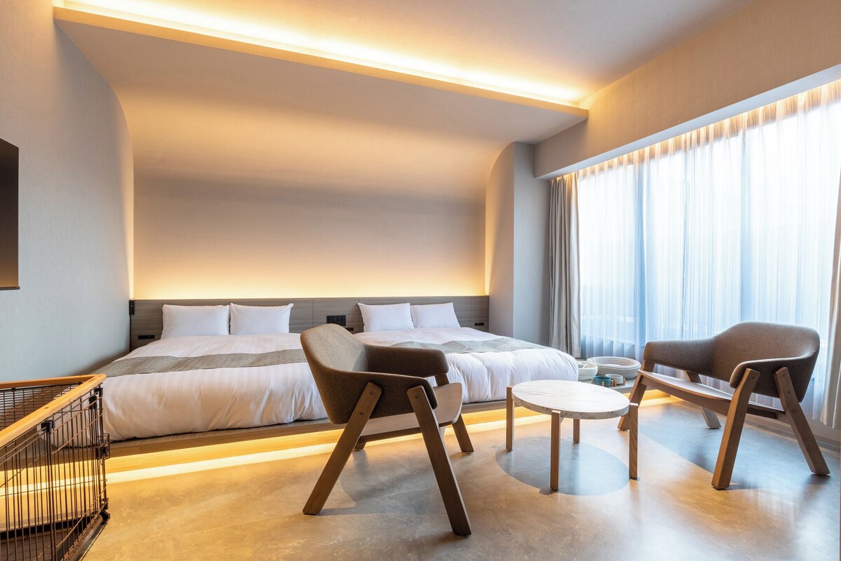 A modern hotel room with a large bed, two armchairs, a small table, and a view of the city through sheer curtains.