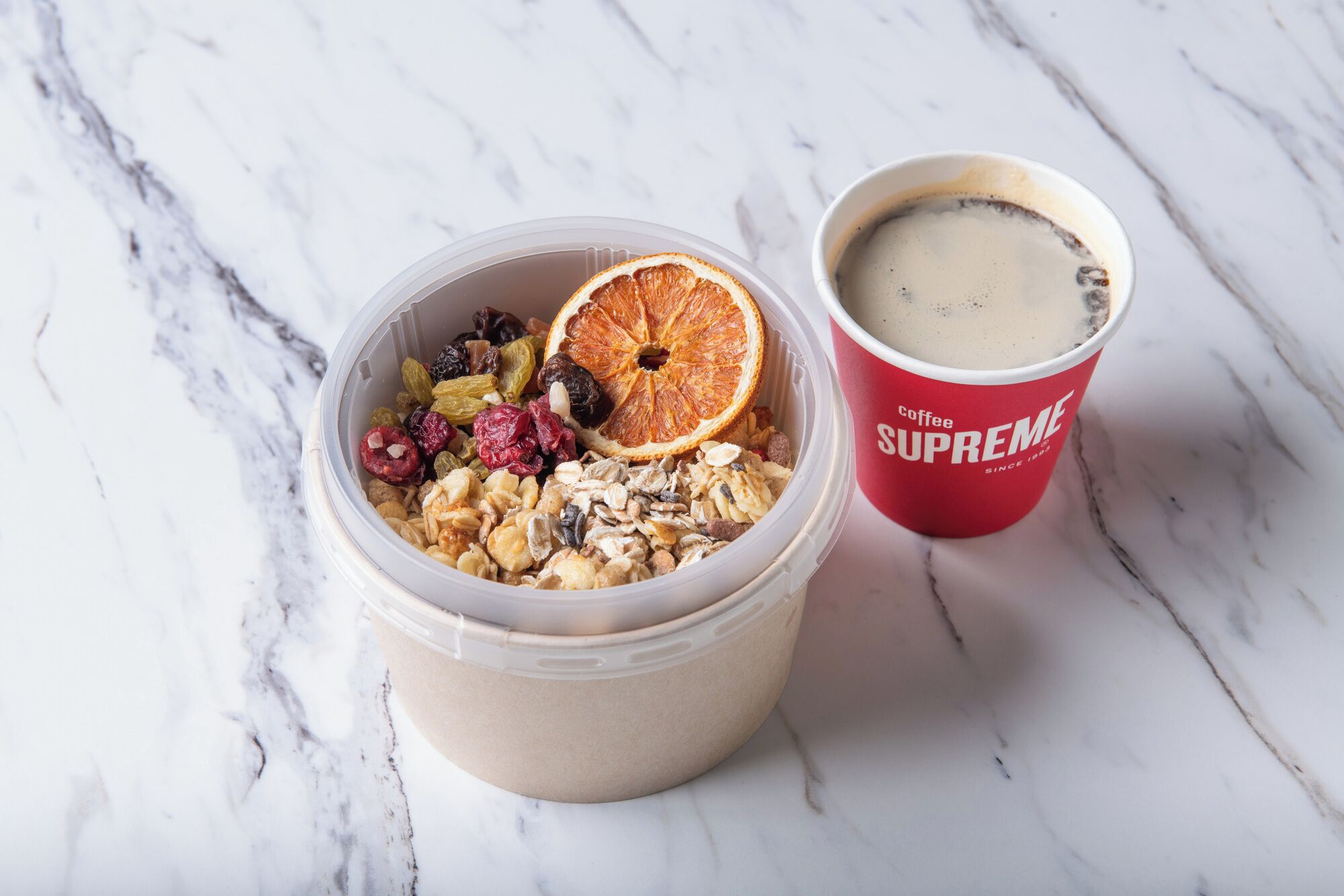 A container of granola with dried fruits and nuts, and a cup of coffee with a red exterior.
