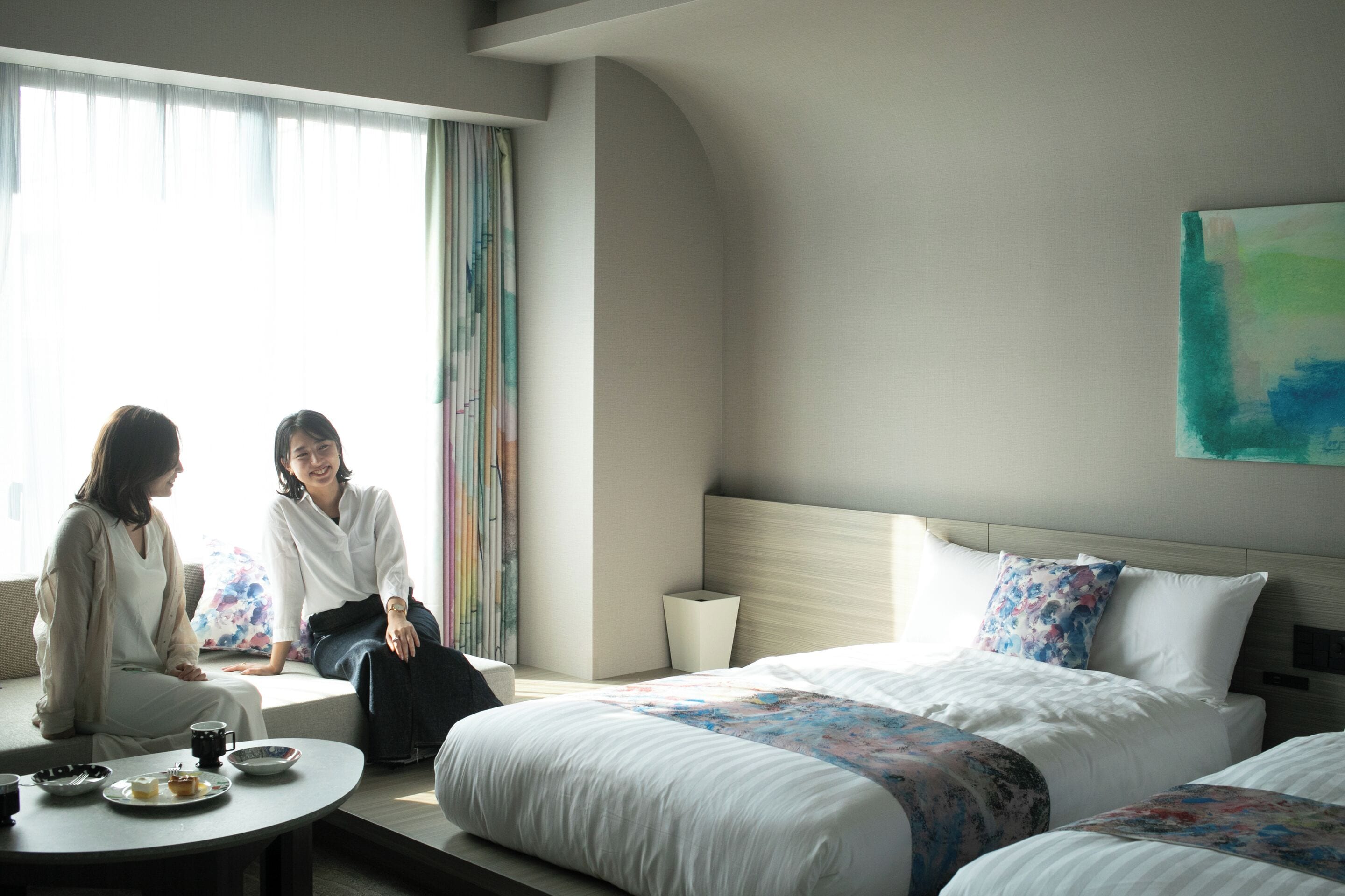 Una habitación de hotel con una cama grande, una cama más pequeña, una mesa redonda con una taza de café y una ventana con cortinas.