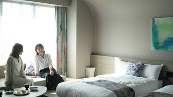 Una habitación de hotel con una cama grande, una cama más pequeña, una mesa redonda con una taza de café y una ventana con cortinas.