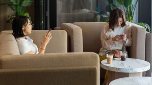 Un ambiente interior moderno con dos personas sentadas en una mesa con bebidas, rodeadas de grandes plantas verdes y un techo de madera.
