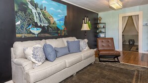 Un salón con un sofá color beige, un sillón marrón, un cuadro grande de una cascada, una lámpara y una estantería.
