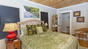 Un dormitorio con una cama grande, una mesita de noche con lámpara, una silla de mimbre y una puerta que da a otra habitación.