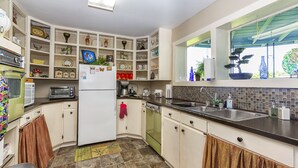 Una cocina compacta con gabinetes blancos, un refrigerador, un microondas y un lavamanos. Hay objetos decorativos en los estantes, y una ventana con vistas a la vegetación exterior.