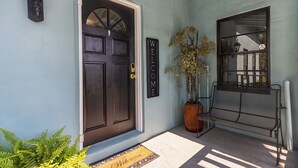 Una puerta de madera oscura con un ventanal semicircular, un letrero que dice 'WELCOME', una planta en maceta, un banco de metal y un felpudo.