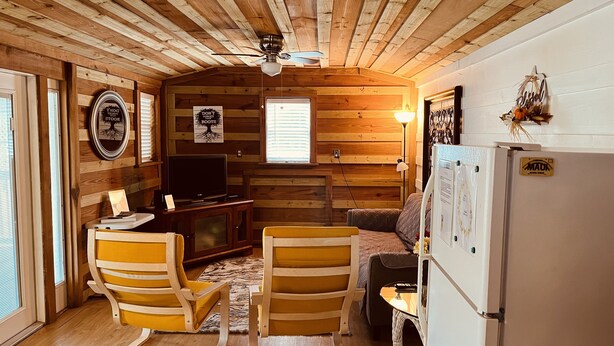 Interior acogedor de una cabaña con paredes de madera, un ventilador de techo, un televisor, dos sillones amarillos, un pequeño sofá y un refrigerador.