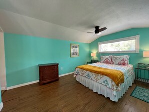 Quarto com paredes verde-água, cômoda de madeira, cama com roupa de cama estampada, ventilador de teto e janela com persianas.