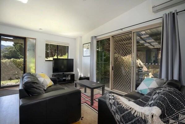 A modern living room with a sofa, a TV, and a table. There is a sliding glass door leading to an outdoor area.