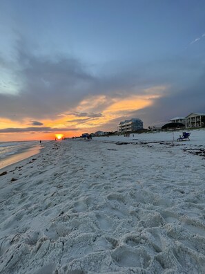 Ein Strand mit Fußabdrücken, einem Sonnenuntergang und Gebäuden im Hintergrund.