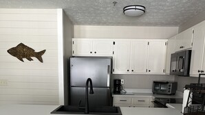 A modern kitchen with white cabinets, a stainless steel refrigerator, a coffee maker, and a toaster. There are four wooden stools at the counter. - Panama City Beach West Side Gem — Steps to the Beach, Poolside, Ground‑Floor (Panama City Beach)