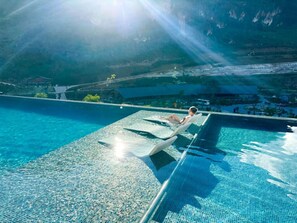 Une piscine à l’eau claire, une personne allongée sur un transat et un paysage montagneux en arrière-plan.