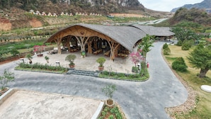 A large, open-air structure with a thatched roof, surrounded by a landscaped garden with potted plants and a paved walkway. - Moc Chau Island - Mountain Park and Resort (Moc Chau)