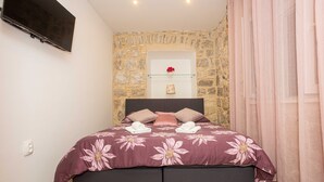 A bedroom with a stone wall, a bed with a floral bedspread, a flat-screen TV, and a glass shelf with a vase. - Apt Sleeps 2 with Sea Proximity (Split)