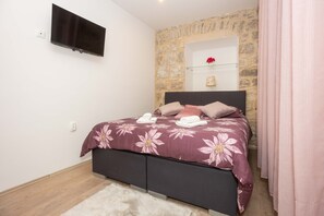 Un dormitorio con pared de piedra, un televisor de pantalla plana, una cama con ropa de cama floral y un pequeño estante con una flor roja.